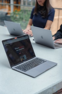 Three people are seated at a long table, each using a laptop. The laptops are Apple MacBooks. One of the laptops has a screen displaying a website with the text 'Where projects run smoothly.' The setting appears to be a modern, well-lit indoor space with greenery visible in the background.