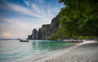 Tropical beach scene featuring traditional wooden boats in calm, turquoise waters alongside a sandy shoreline. Lush green foliage and steep limestone cliffs provide a dramatic backdrop, while clouds scatter across the blue sky.