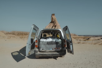 A van with open back doors is parked in a desert landscape. The interior is visible, filled with a covered bed or sleeping area, suggesting a travel or camping setup. The background features barren, rocky land and clear skies.