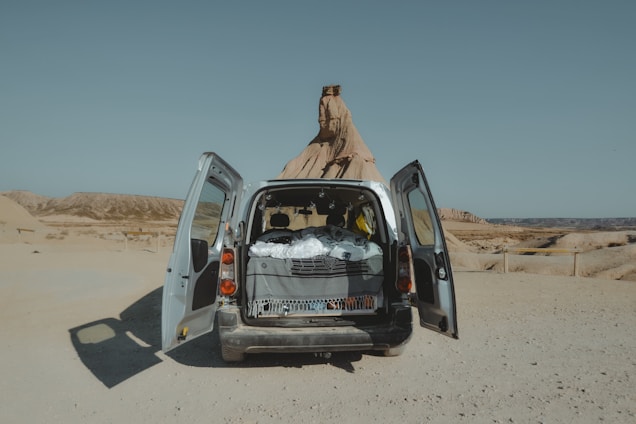 A van with open back doors is parked in a desert landscape. The interior is visible, filled with a covered bed or sleeping area, suggesting a travel or camping setup. The background features barren, rocky land and clear skies.