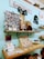 A brightly lit, cozy shop display featuring various handmade items and decorations. A wooden shelf mounted on a light blue wall holds a variety of items including a cluster of photographs, a small pink camera, potted plants, and wooden decor. Below this shelf, a wooden counter displays an assortment of small merchandise such as cards, stickers, earrings, and other knick-knacks. A sign on the wall reads 'Happiness is homemade,' conveying a welcoming and homey atmosphere.