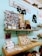 A brightly lit, cozy shop display featuring various handmade items and decorations. A wooden shelf mounted on a light blue wall holds a variety of items including a cluster of photographs, a small pink camera, potted plants, and wooden decor. Below this shelf, a wooden counter displays an assortment of small merchandise such as cards, stickers, earrings, and other knick-knacks. A sign on the wall reads 'Happiness is homemade,' conveying a welcoming and homey atmosphere.