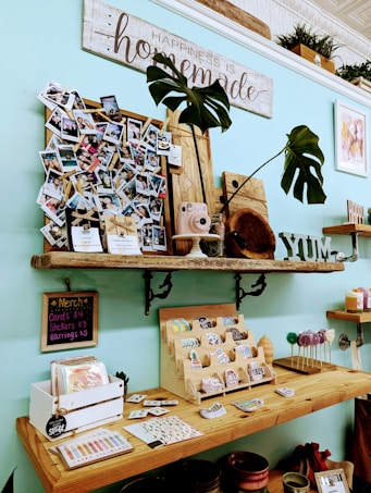 A brightly lit, cozy shop display featuring various handmade items and decorations. A wooden shelf mounted on a light blue wall holds a variety of items including a cluster of photographs, a small pink camera, potted plants, and wooden decor. Below this shelf, a wooden counter displays an assortment of small merchandise such as cards, stickers, earrings, and other knick-knacks. A sign on the wall reads 'Happiness is homemade,' conveying a welcoming and homey atmosphere.
