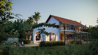 A two-story house with an arched entrance and a red tiled roof, surrounded by lush greenery and tall palm trees. A vintage scooter and an old green Land Rover are parked nearby. Warm interior lights illuminate the house, creating a cozy atmosphere.