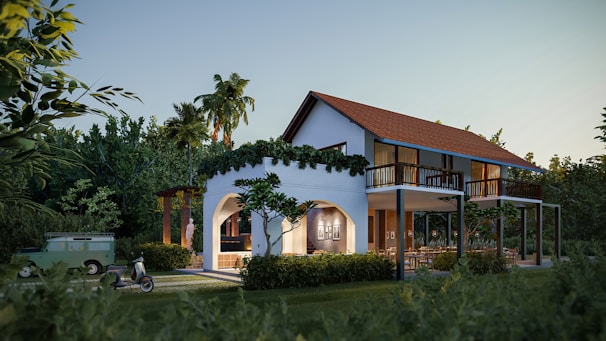 A two-story house with an arched entrance and a red tiled roof, surrounded by lush greenery and tall palm trees. A vintage scooter and an old green Land Rover are parked nearby. Warm interior lights illuminate the house, creating a cozy atmosphere.