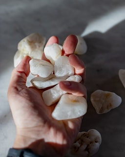 A hand holds a collection of white stones, with more stones scattered on a gray surface. Soft shadows and light create a calm and natural atmosphere.