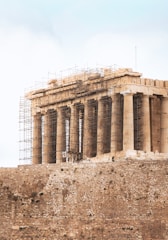 A historical ancient Greek temple stands on a rocky outcrop, surrounded by scaffolding as it undergoes restoration. The structure is characterized by its classical columns and weathered stone, conveying a sense of antiquity and architectural grandeur.