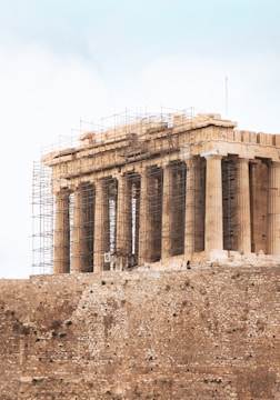A historical ancient Greek temple stands on a rocky outcrop, surrounded by scaffolding as it undergoes restoration. The structure is characterized by its classical columns and weathered stone, conveying a sense of antiquity and architectural grandeur.