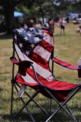 A folding chair is prominently displayed with a fabric design resembling the American flag, featuring stars and stripes. The chair is situated on a grassy field with people and trees in the blurred background, suggesting an outdoor gathering or event.