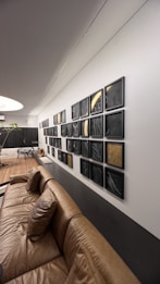 The modern interior features a leather couch positioned against a wall adorned with a grid of abstract art pieces in black frames. The artworks include various geometric patterns and one with a gold accent. Natural light illuminates the space, and a potted plant is visible alongside other sleek, contemporary furniture.