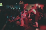 A performer on stage interacts energetically with an enthusiastic audience. People are reaching out towards the performer, creating a lively and engaged atmosphere. The scene is dimly lit with bright spotlights and scattered colored lighting, suggesting a vibrant and dynamic live performance.