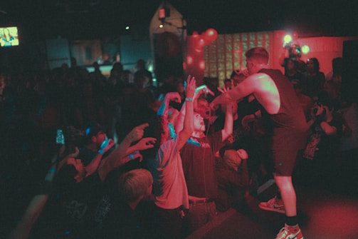 A performer on stage interacts energetically with an enthusiastic audience. People are reaching out towards the performer, creating a lively and engaged atmosphere. The scene is dimly lit with bright spotlights and scattered colored lighting, suggesting a vibrant and dynamic live performance.