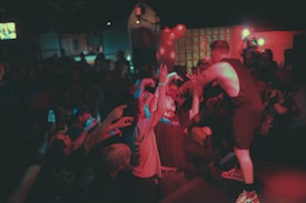 A performer on stage interacts energetically with an enthusiastic audience. People are reaching out towards the performer, creating a lively and engaged atmosphere. The scene is dimly lit with bright spotlights and scattered colored lighting, suggesting a vibrant and dynamic live performance.