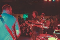 A band is performing on stage with several musicians visible, including a guitarist and a drummer. The audience can be seen in the background, enjoying the live music. The lighting casts colorful hues over the scene, creating a vibrant atmosphere.