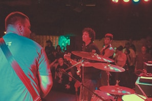 A band is performing on stage with several musicians visible, including a guitarist and a drummer. The audience can be seen in the background, enjoying the live music. The lighting casts colorful hues over the scene, creating a vibrant atmosphere.
