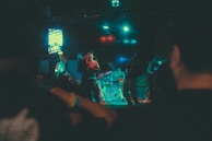 A dimly lit concert scene captures a band performing on stage with several musicians playing instruments like guitars and drums. The atmosphere is vibrant with colorful lighting, creating an engaging visual experience. The audience is visible, partially silhouetted in the foreground.