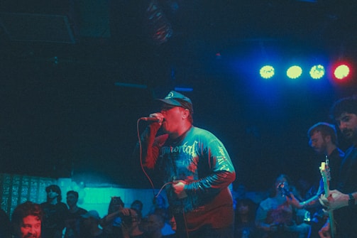 A musician passionately sings into a microphone on stage, wearing a hat and long-sleeved shirt. Band members are playing instruments nearby, and an audience is visible in the dimly lit background. The lighting features blue and red hues, creating a vibrant atmosphere.