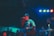 A musician passionately sings into a microphone on stage, wearing a hat and long-sleeved shirt. Band members are playing instruments nearby, and an audience is visible in the dimly lit background. The lighting features blue and red hues, creating a vibrant atmosphere.