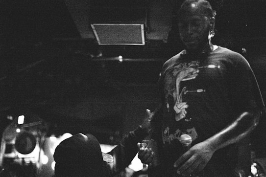 A black and white image capturing a person in a casual t-shirt holding a microphone. The scene appears dimly lit, suggesting a nocturnal atmosphere, possibly in a club or a performance venue. Another individual, wearing a hat, is partially visible, reaching out toward the person with the microphone.