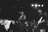 A black and white image capturing a live music performance in a dimly lit venue. The scene includes a singer passionately holding a microphone and wearing casual attire, while a guitarist is playing beside him. A crowd of spectators appears in the background, enjoying the music. Some balloons are visible in a corner.