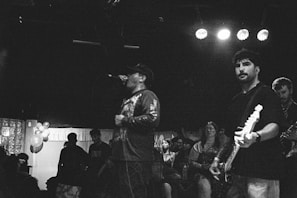 A black and white image capturing a live music performance in a dimly lit venue. The scene includes a singer passionately holding a microphone and wearing casual attire, while a guitarist is playing beside him. A crowd of spectators appears in the background, enjoying the music. Some balloons are visible in a corner.
