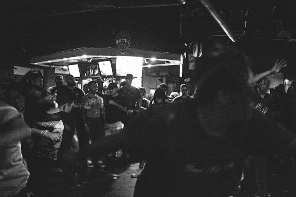 A black and white image capturing a crowded nightclub or music venue. People are gathered closely together, with some individuals dancing or moving energetically. The atmosphere appears vibrant and lively, with dim lighting enhancing the scene's intensity. Multiple screens are visible in the background, possibly displaying visuals or information relevant to the event.