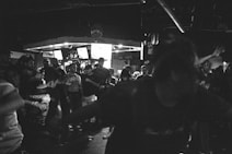 A black and white image capturing a crowded nightclub or music venue. People are gathered closely together, with some individuals dancing or moving energetically. The atmosphere appears vibrant and lively, with dim lighting enhancing the scene's intensity. Multiple screens are visible in the background, possibly displaying visuals or information relevant to the event.