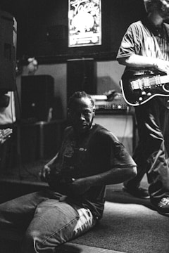 A black and white photograph captures a man sitting on the floor with a casual posture, surrounded by musical equipment. Another person is standing nearby, holding an electric guitar. The setting appears to be indoors, possibly at a live music venue or rehearsal space, with dim lighting contributing to the moody atmosphere.