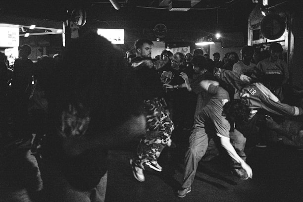 A lively scene with a large group of people gathered in a dimly lit environment, likely a club or concert setting. Individuals are engaged in energetic dancing or movement, capturing a moment of intense activity and interaction.