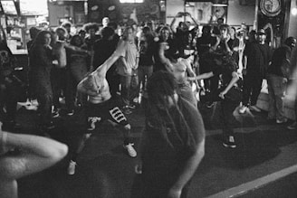 A lively crowd gathers in a dimly lit indoor venue. The focus is on a group of people energetically dancing in the foreground, with one individual prominently leaning forward. The scene is captured in black and white, adding a nostalgic and intense feel.