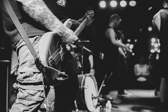A black and white photograph captures a live band performance. A tattooed musician plays an electric guitar on stage with other band members visible in the background. The image has a dynamic, candid feel, with stage lights and a subtle audience present.