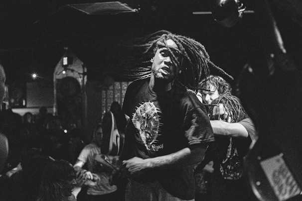 A person with long, flowing dreads is energetically moving in a dimly lit venue. The scene appears chaotic, with a crowd in the background adding to the dynamic atmosphere. The image is in black and white, emphasizing contrast and movement.