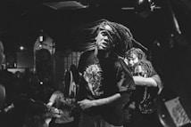 A person with long, flowing dreads is energetically moving in a dimly lit venue. The scene appears chaotic, with a crowd in the background adding to the dynamic atmosphere. The image is in black and white, emphasizing contrast and movement.