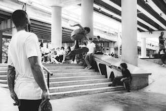 A group of young people gather at a skatepark under a highway overpass. One skateboarder jumps off a set of stairs while others watch. Another person is sitting on the ledge taking a photo or video. The environment is urban and energetic, with visible skateboards and bystanders.