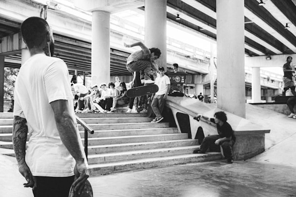 A group of young people gather at a skatepark under a highway overpass. One skateboarder jumps off a set of stairs while others watch. Another person is sitting on the ledge taking a photo or video. The environment is urban and energetic, with visible skateboards and bystanders.