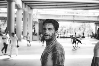 A black and white photograph depicting a tattooed man in the foreground under a bridge. The setting has a gritty urban feel with people in the background, some of whom appear to be skating. The man is shirtless and looking towards the camera, while others are casually dispersed throughout the scene.