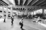 A group of skateboarders is gathered under a highway overpass, engaging in various tricks and maneuvers. The scene is lively with several people spectating in the background. The area is a large open space with concrete pillars supporting the overhead structure.