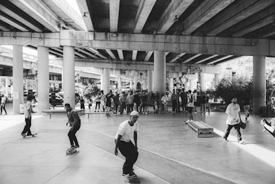 A group of skateboarders is gathered under a highway overpass, engaging in various tricks and maneuvers. The scene is lively with several people spectating in the background. The area is a large open space with concrete pillars supporting the overhead structure.