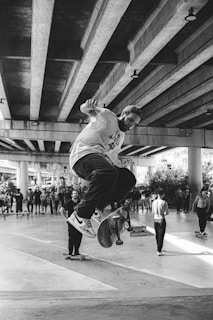 A black and white image of a skateboarder performing a trick under a large concrete bridge. The scene includes a group of spectators and other skateboarders watching the trick. The concrete beams overhead create a pattern and the area appears to be an urban skate park.