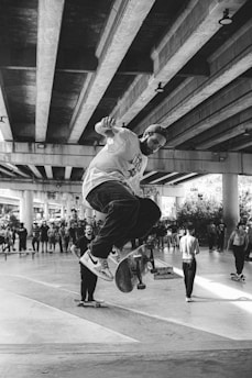 A black and white image of a skateboarder performing a trick under a large concrete bridge. The scene includes a group of spectators and other skateboarders watching the trick. The concrete beams overhead create a pattern and the area appears to be an urban skate park.