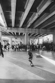 A skateboarder in motion under a large concrete overpass, observed by a group of spectators in the background. The setting has an urban and industrial vibe, with concrete pillars and shadows cast by the overhead structure.