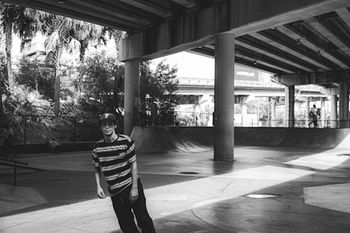 A skateboarder is skating under an urban overpass. The area is shaded with concrete structures and surrounded by greenery.