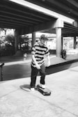 A black and white scene of a skate park beneath a highway overpass. A young man in a striped shirt and cap is skateboarding towards the camera. Several other people are seen in the background, engaging in similar activities. The overpass structure casts shadows on the concrete surface, adding to the urban atmosphere.