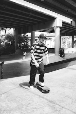 A black and white scene of a skate park beneath a highway overpass. A young man in a striped shirt and cap is skateboarding towards the camera. Several other people are seen in the background, engaging in similar activities. The overpass structure casts shadows on the concrete surface, adding to the urban atmosphere.