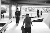 A black and white photo of a skatepark under a bridge, with several people skateboarding. Two individuals in the foreground are walking towards the camera, one holding a skateboard. Others can be seen in the background on ramps and walkways. The area is surrounded by concrete structures and graffiti.