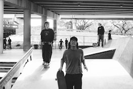 A black and white photo of a skatepark under a bridge, with several people skateboarding. Two individuals in the foreground are walking towards the camera, one holding a skateboard. Others can be seen in the background on ramps and walkways. The area is surrounded by concrete structures and graffiti.