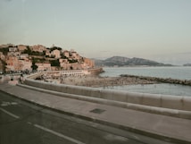 A coastal cityscape with a blend of urban and natural elements. Houses with colorful facades are perched on a sloping hillside overlooking a bustling beachfront. The sandy beach is dotted with people enjoying leisure activities alongside a calm sea. A stone jetty extends into the ocean, while mountains rise majestically in the background under a clear sky. A road with a curved barrier runs adjacent to the beach.