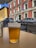 A glass of beer is placed on a wooden table at an outdoor seating area, with a street view in the background featuring parked cars and a red building with multiple windows and flags.