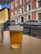 A glass of beer is placed on a wooden table at an outdoor seating area, with a street view in the background featuring parked cars and a red building with multiple windows and flags.