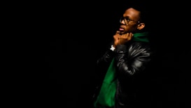 A person wearing a dark leather jacket and a green hoodie stands against a black background, adjusting the hood. They are wearing glasses and a watch, and their facial expression is contemplative.
